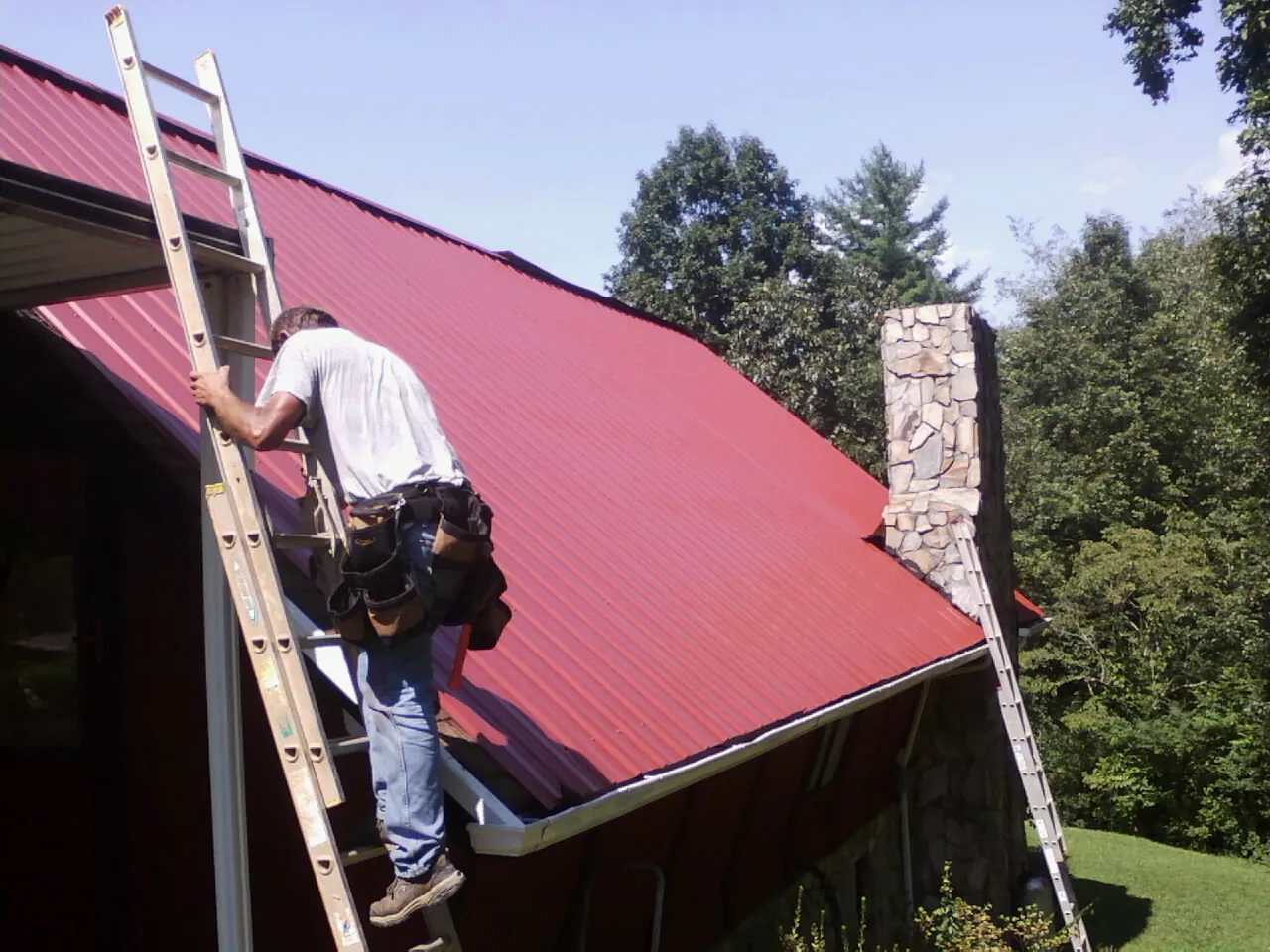 Roofer on a ladder beside a red metal roof and stone chimney — Soffit & Fascia Repair by Roofing Brothers in Pine Meadow